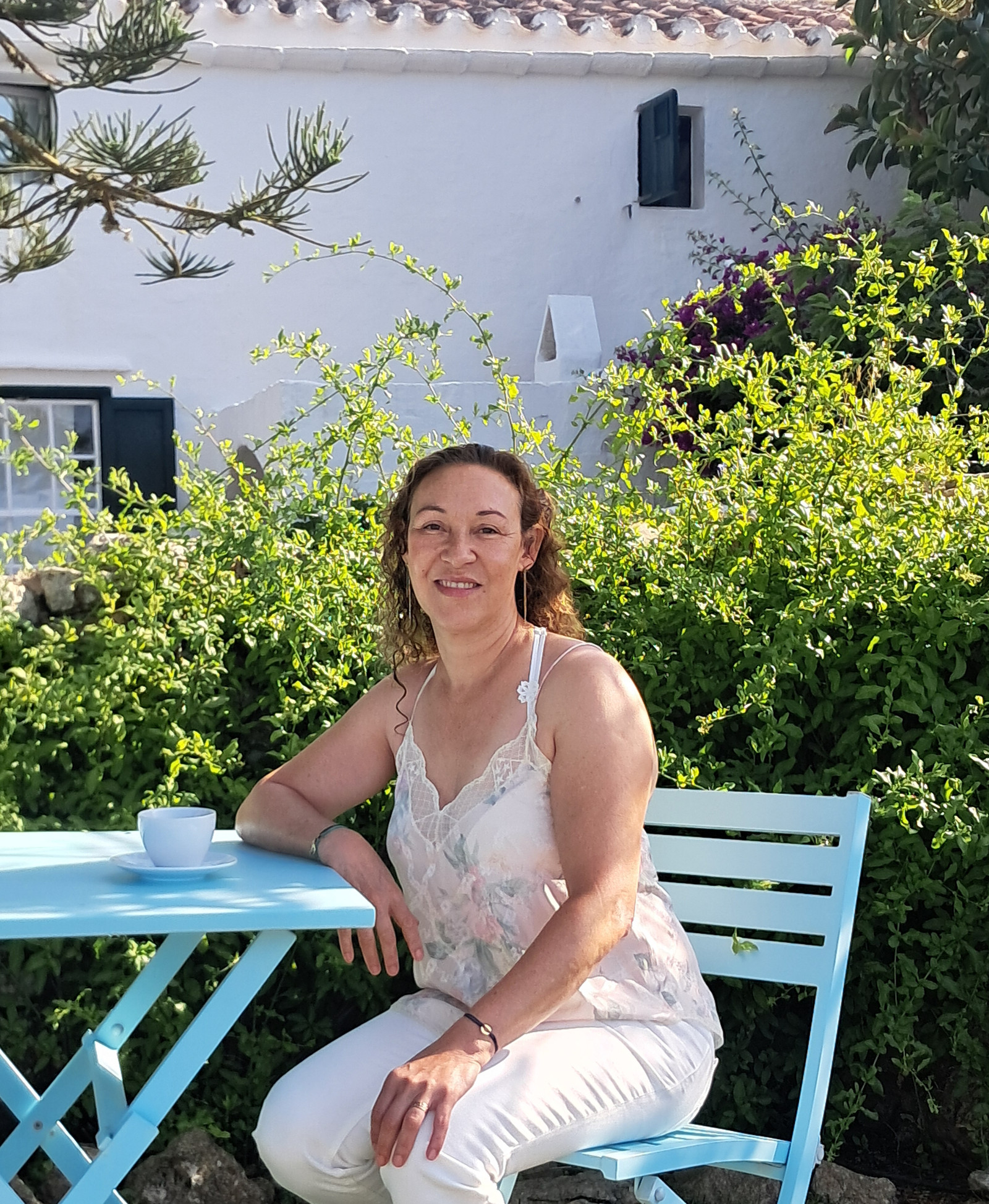 Marjolaine Sami assise à une table de jardin devant une maison blanche aux volets sombres